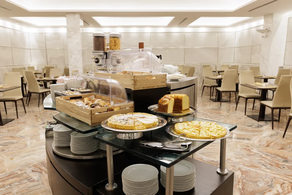 Nell'immagine una parte del buffet della prima colazione: tre tipologie di torte e brioche. Intorno al tavolo del buffet i tavolini apparecchiati per la prima colazione.