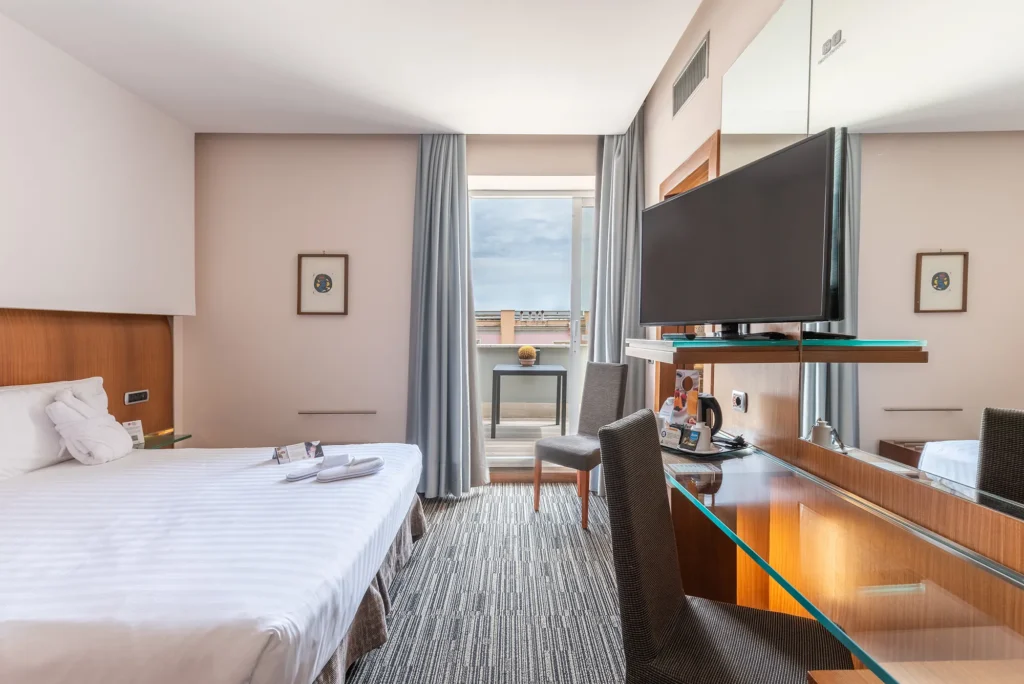 Nell'immagine si vede il letto matrimoniale con di fronte una scrivania con una sedia e sopra una mensola con la televisione. Di lato una portafinestra che consente di accedere al balcone.