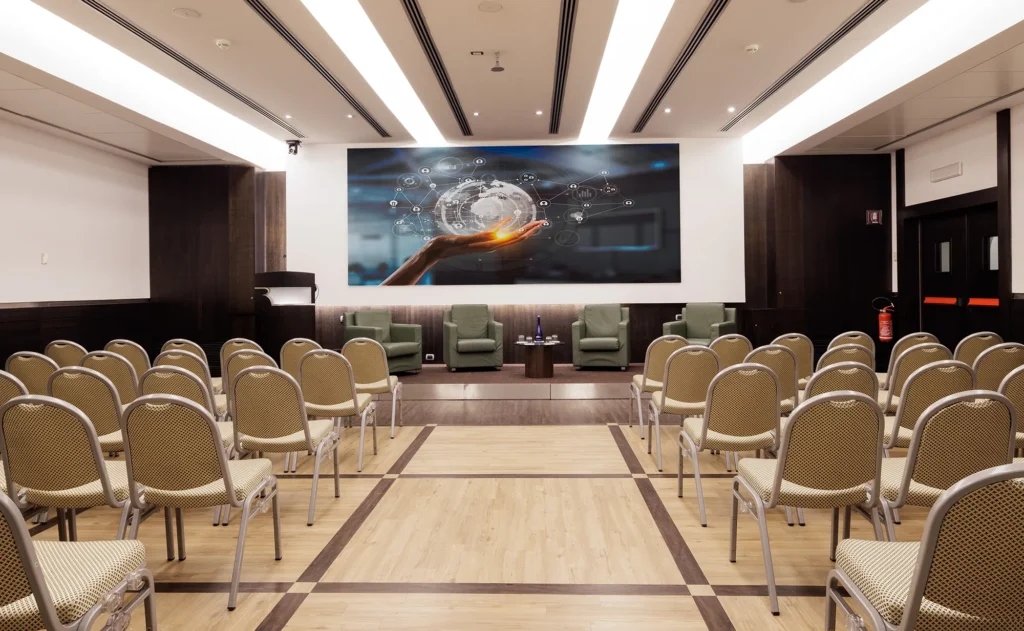 Nell'immagine la Sala Meeting Anna con disposizione a platea. Di fronte alla platea un palco con quattro poltrone per i relatori e un tavolino al centro con delle bottiglie d'acqua.