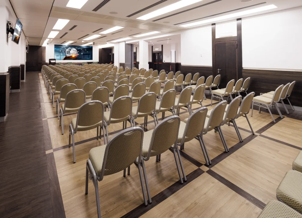 Nell'immagine la Sala Meeting AnnaMaria con disposizione a platea. Di fronte alla platea un palco con quattro poltrone per i relatori e un tavolino al centro con delle bottiglie d'acqua.
