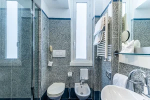 Nell'immagine il bagno con doccia fronte il lavandino. Sopra uno specchio rettangolare con una mensola in vetro. Di lato i sanitari e una finestra.